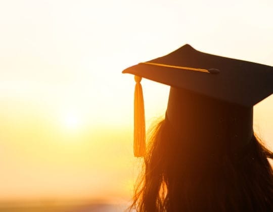 Graduate wears a black cap with yellow tassel, facing away from camera and looking at a sunset.