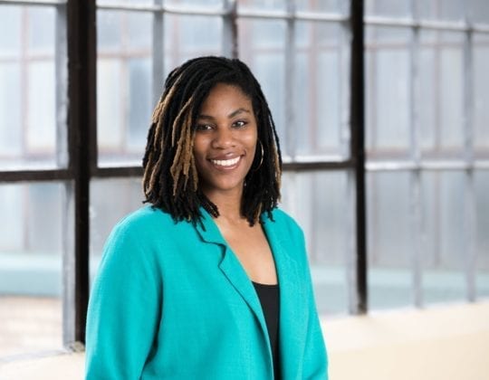 Portrait photo of Mya Williams, Kenzie Academy alum and UX Engineering facilitator, smiling and standing in front of sunny windows.