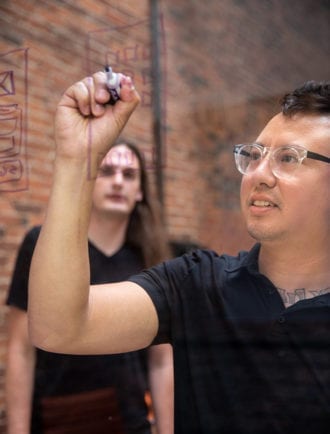 Man with glasses drawing UX design on a clear board with man watching in background.