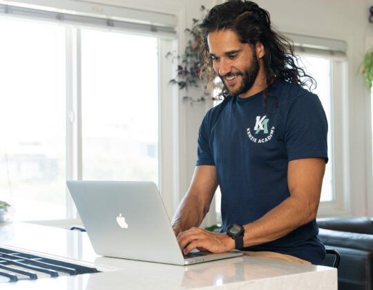 Man with long hair typing on laptop wearing a blue Kenzie Academy tshirt